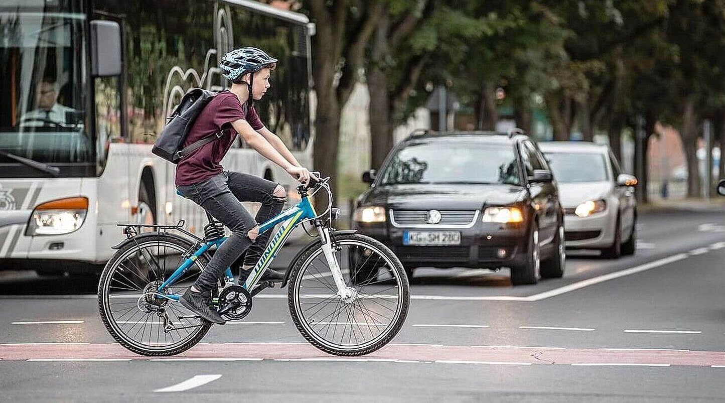 Junge fährt auf dem Fahrrad über einen rot markierten Radweg an wartenden Autos vorbei.